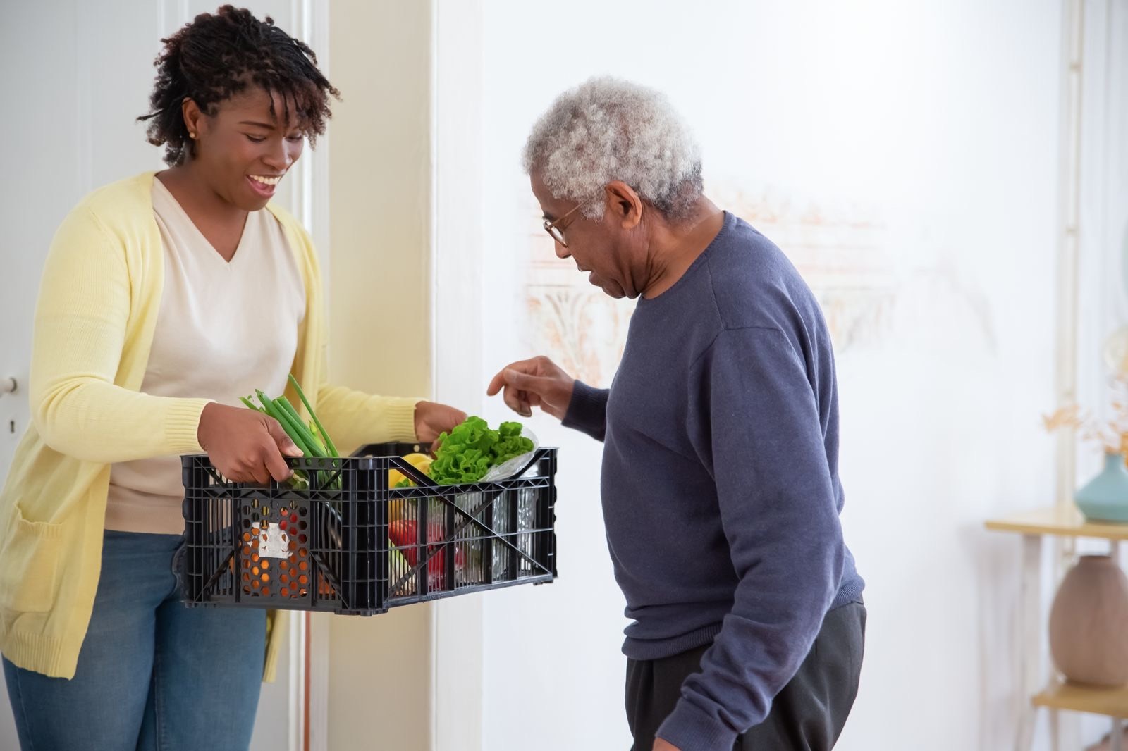 A woman brings a crate of fresh vegetables to an older man in his home.
